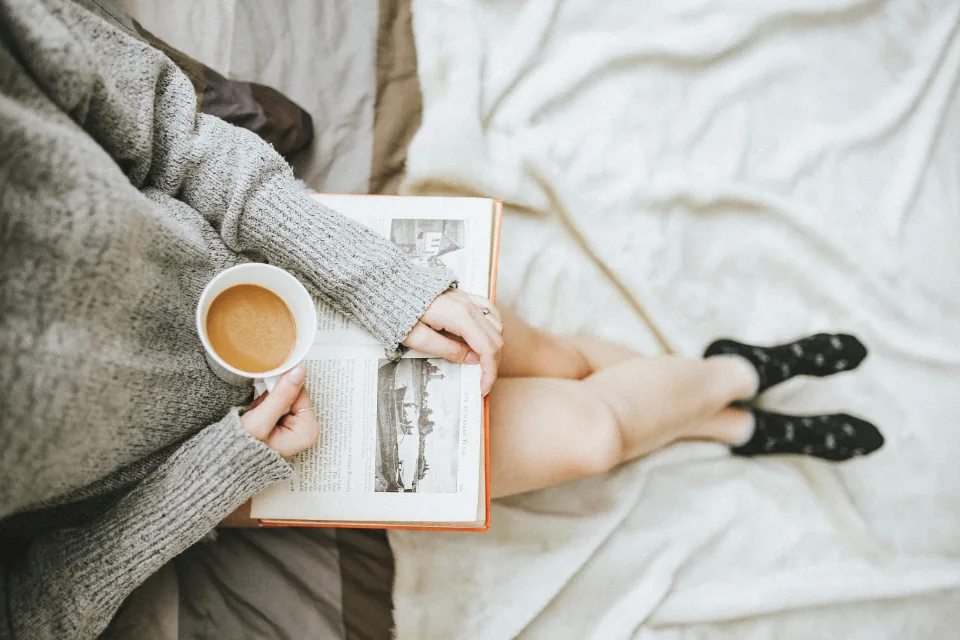 A woman curled up on a bed under a soft knit blanket with a book and a cup of coffee, winding down with calming affirmations