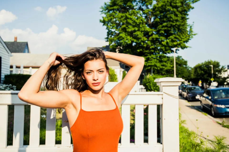 A young woman in an orange top standing in the sun with her hands in her hair, relaxed and self-assured