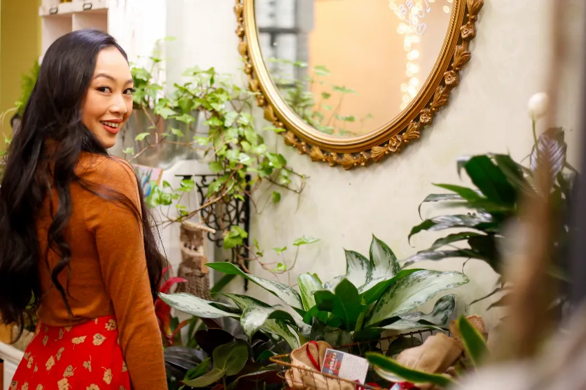 A woman in a rust-coloured sweater smiling over her shoulder beside a gold mirror and plants