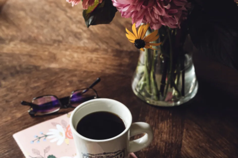 A cup of coffee on a floral notebook beside a vase of flowers on a wooden table