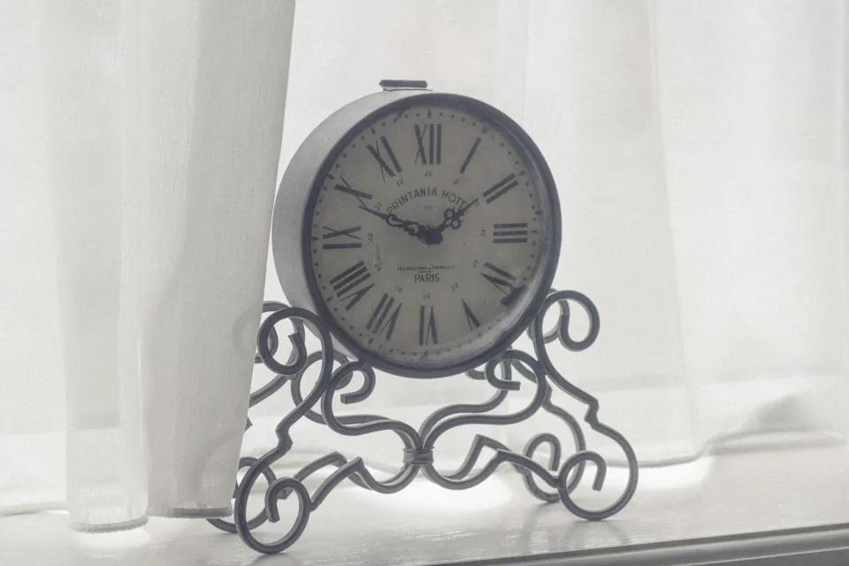 A vintage clock on a windowsill behind a white curtain, a quiet reminder that manifesting takes time