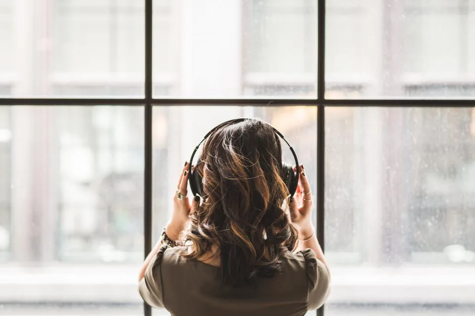 A woman with headphones on, standing at a tall window, listening to affirmations