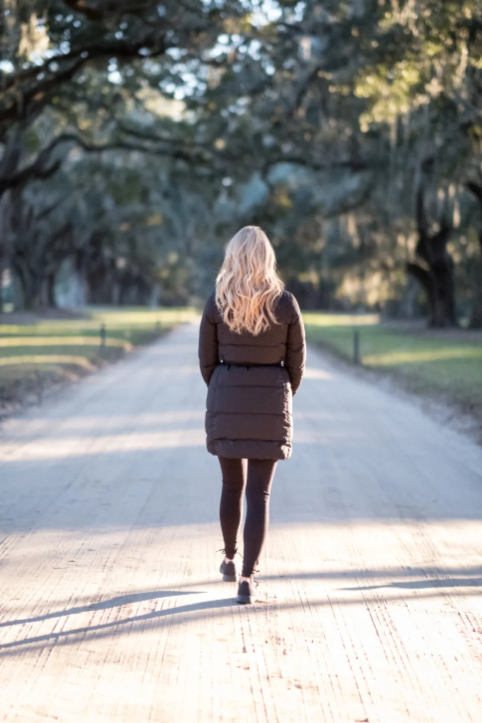 A woman walking down a tree-lined road in soft evening light, taking a step toward her goal