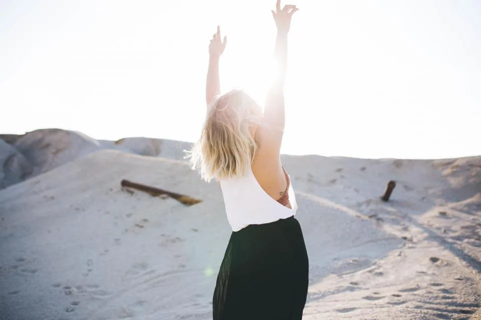 A woman with blonde hair raising her arms in bright sunlight, feeling free in the life she is manifesting