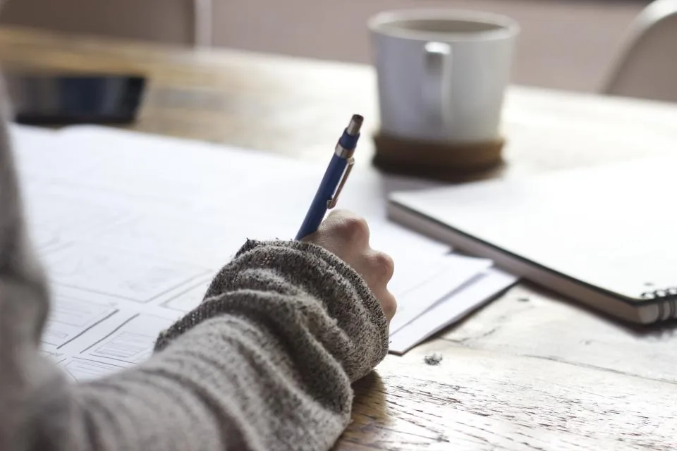 A woman in a soft grey sweater writing her own affirmations at a wooden table with a cup of tea