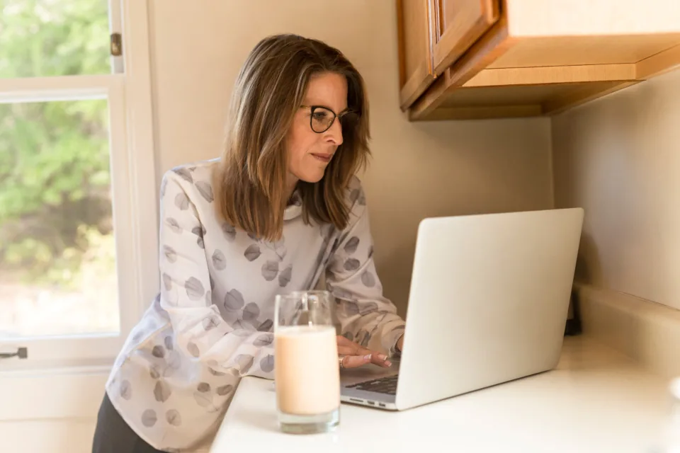 A woman in glasses preparing at her laptop in a sunny kitchen