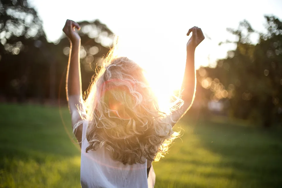 A woman raising her arms into warm evening sunlight in a park, a feeling the law of attraction is often linked to