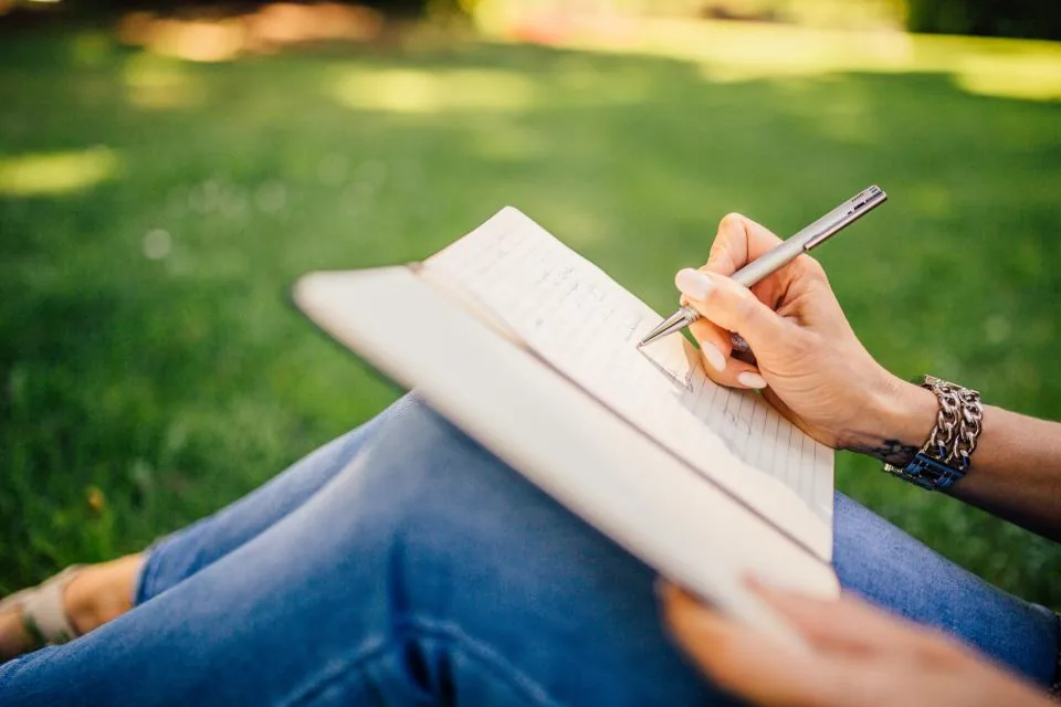 A woman sitting on the grass writing a list in an open notebook, a simple way to start with the law of attraction