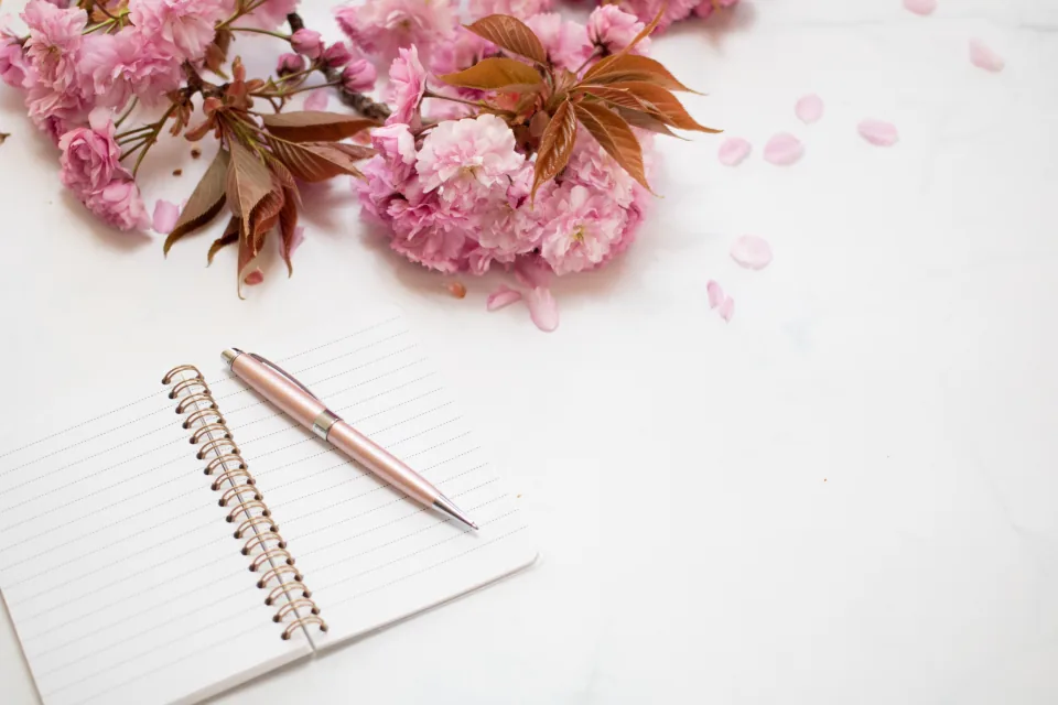 An open notebook and a rose-gold pen on a white desk with pink cherry blossoms