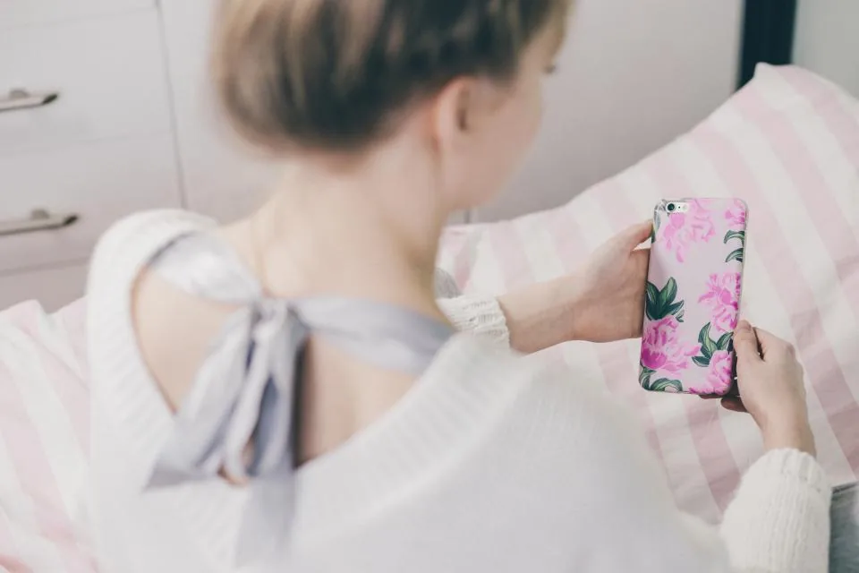 A woman in a soft white sweater reading her morning affirmations on her phone in a blush-pink bedroom