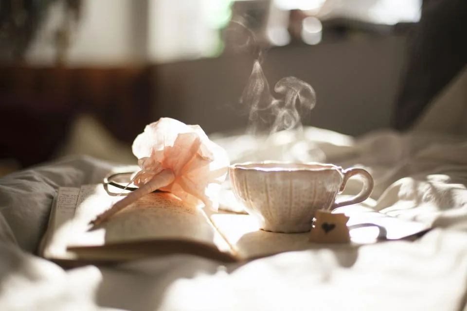 A cup of steaming tea and an open book on a bed in soft morning sunlight