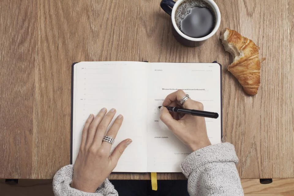 A woman in a cozy sweater writing a manifestation script in a journal with coffee and a croissant