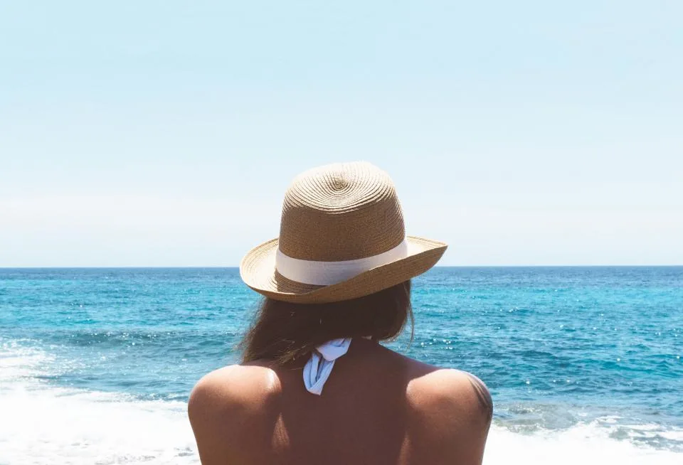 A woman in a straw hat looking out at a bright blue sea, picturing a trip she wants to manifest